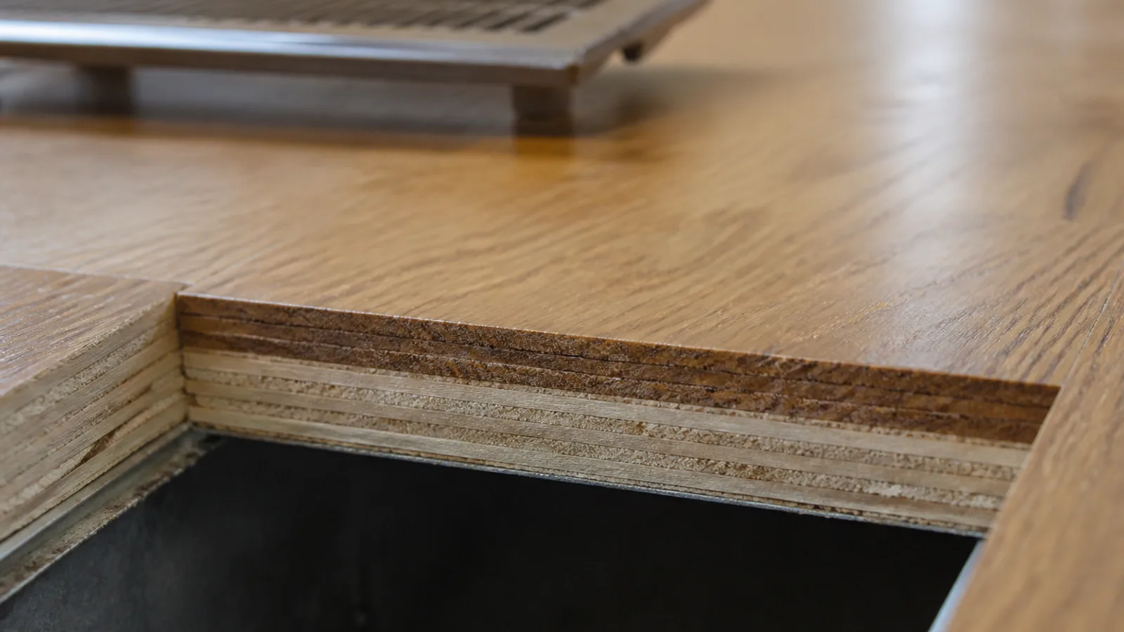 Open floor heating register in an engineered oak floor, exposing the edges of the boards around it