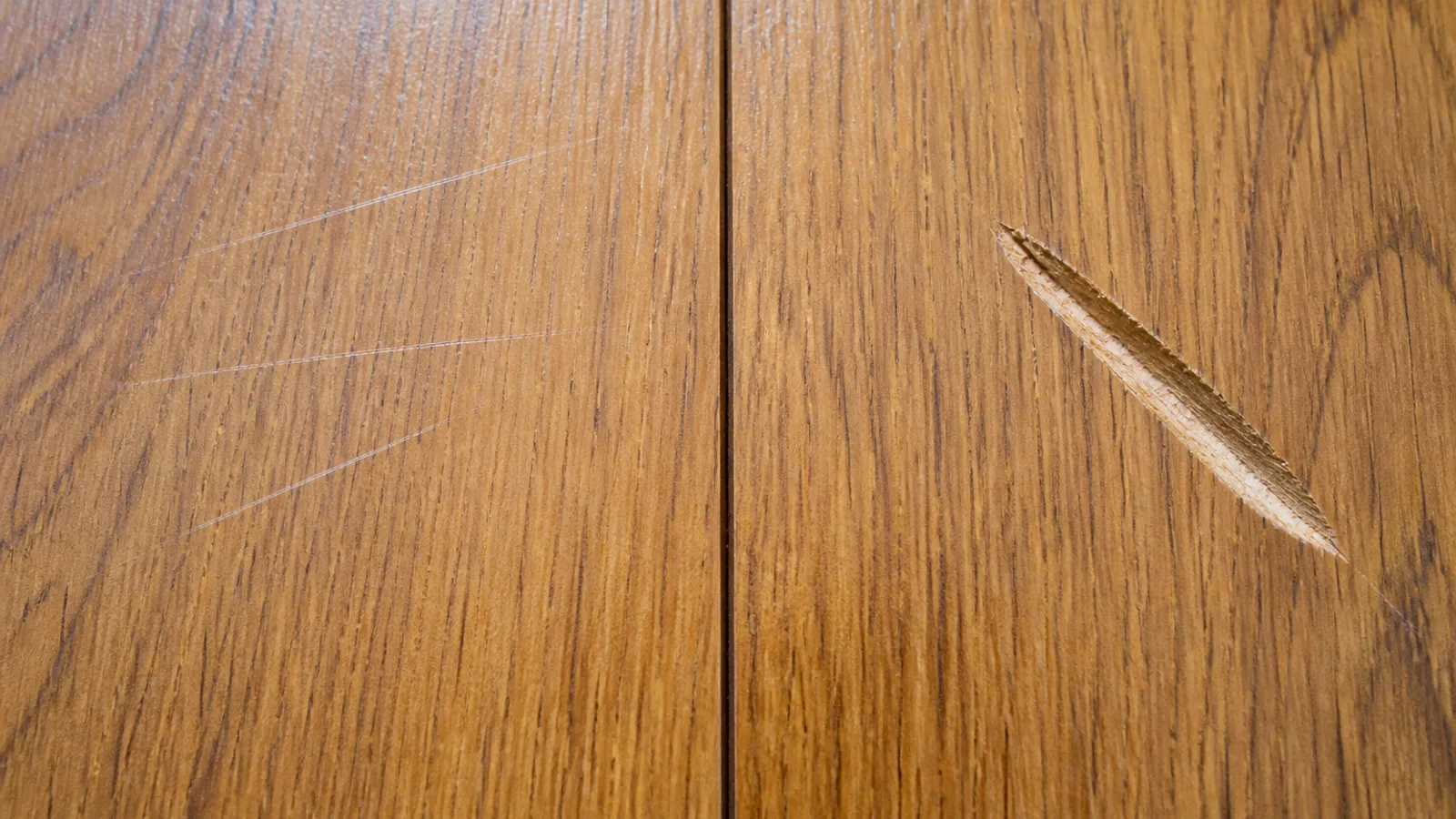 Extreme close-up of oak floorboards showing shallow pale scuff marks sitting on the glossy surface beside a deeper gouge that has cut through into the raw wood