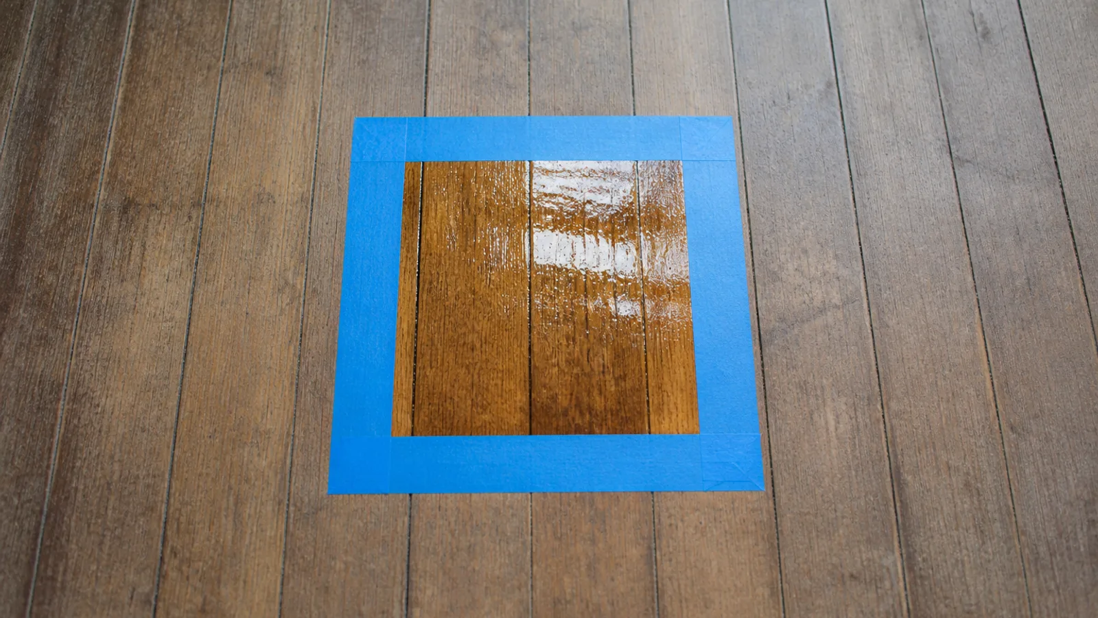 A small square of fresh glossy clear finish applied as a test patch on a dull worn wood floor, its edges masked off with painter's tape