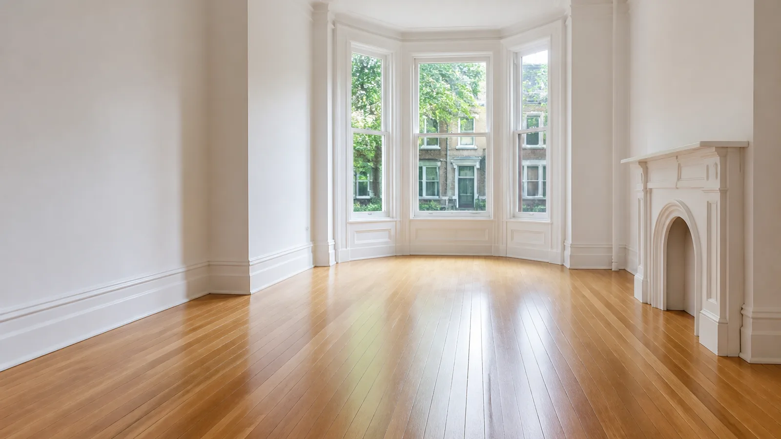 An empty period row-house front room with a freshly refinished golden oak floor running toward a tall bay window, white walls and baseboards